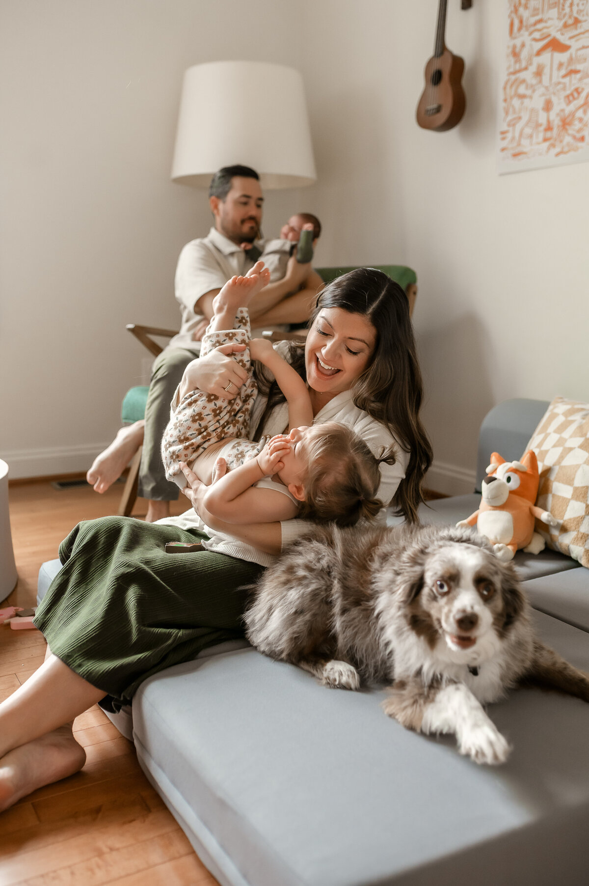 Mother plays with oldest daughter while dog sits comfortably, and dad is sitting in a chair behind them, holding the baby Ijamsville MD