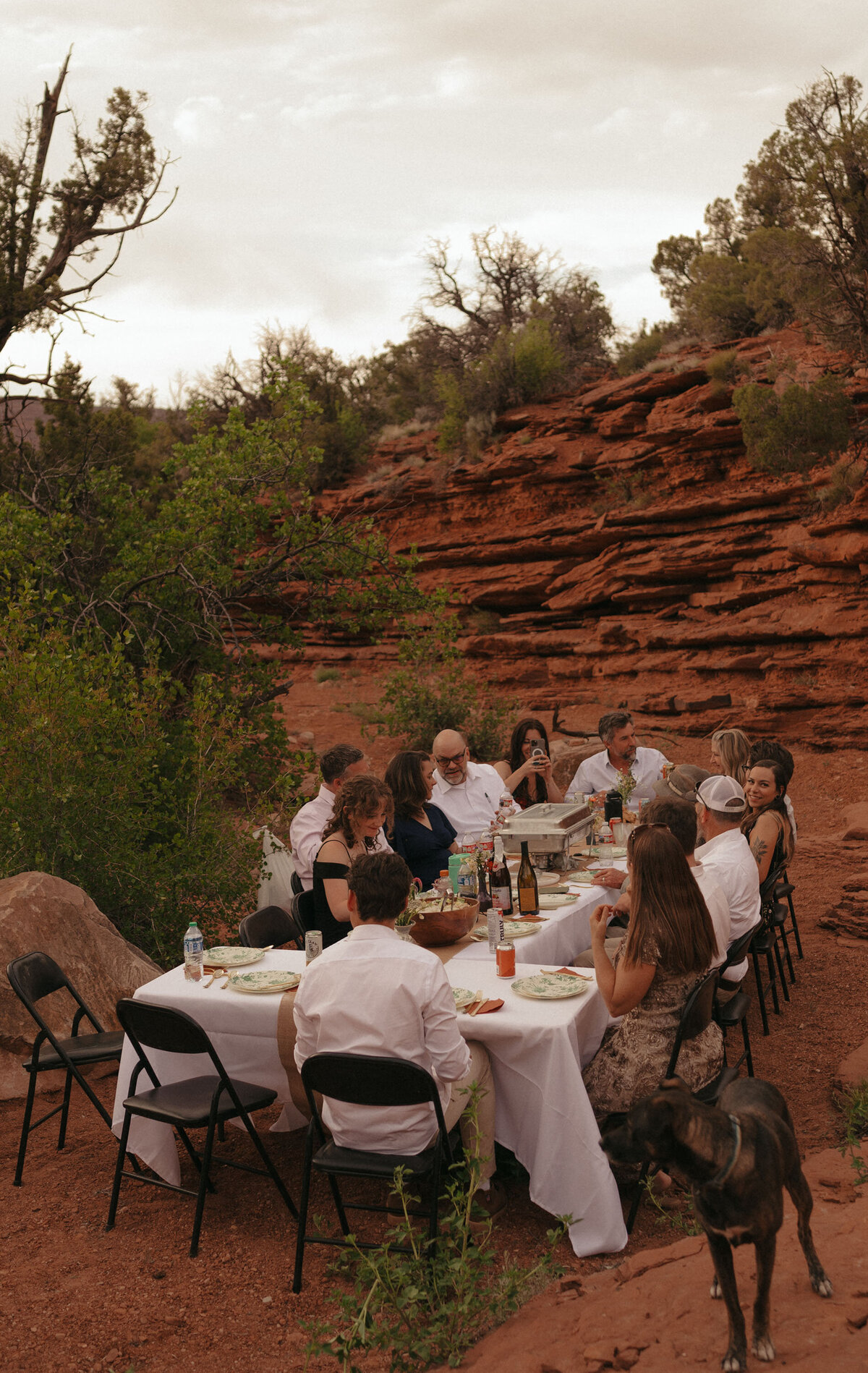 family members at a wedding sit at a table and enjoy a meal against a red sandstone background in the Moab, Utah desert