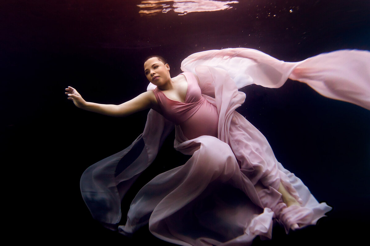 An exquisite underwater maternity photo captured by a Dallas underwater maternity photographer, featuring a woman floating serenely against a dark background. Her flowing pink gown swirls gracefully around her as she extends one arm outward, creating a sense of gentle motion. This Dallas underwater maternity photoshoot emphasizes elegance and tranquility in a beautifully composed scene.