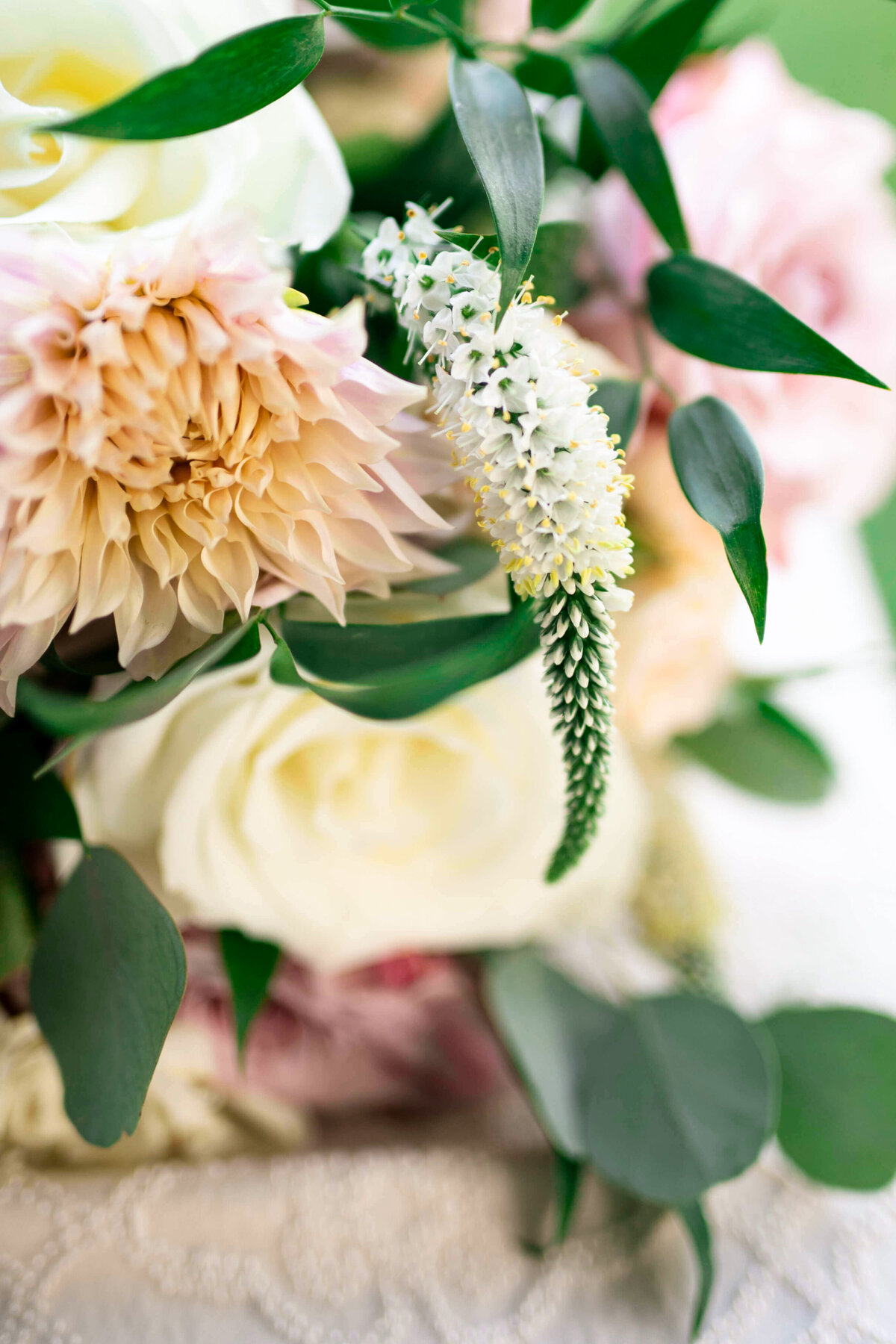 A close-up of a floral arrangement featuring pale pink and cream flowers, including roses and dahlias, accented with green leaves and a white veronica flower, on a lace surface.
