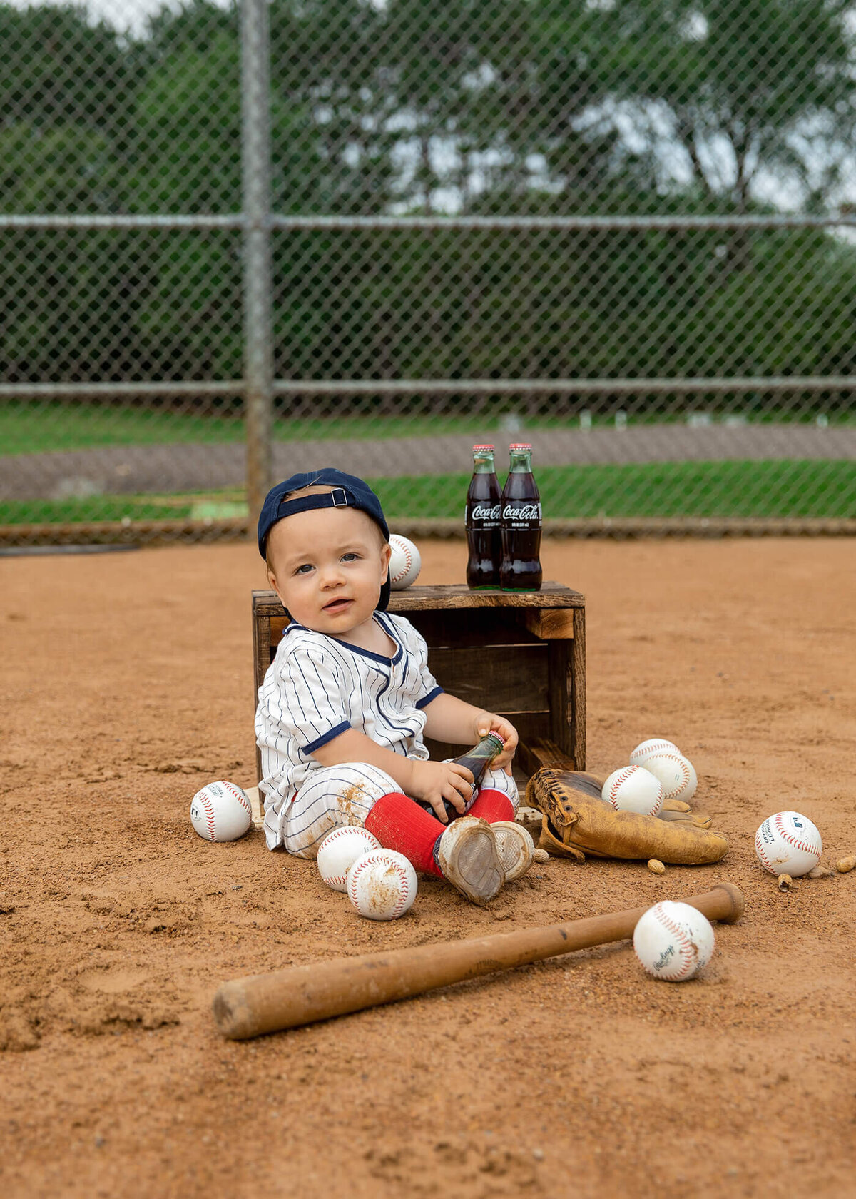 1 year old boy sitting on a baseball diamond with balls, bat and glove sitting around him.