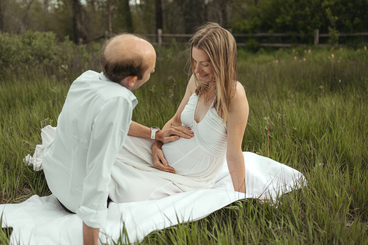 pregnant mom and dad sitting together on white blanket holding and looking at belly in firestone colorado