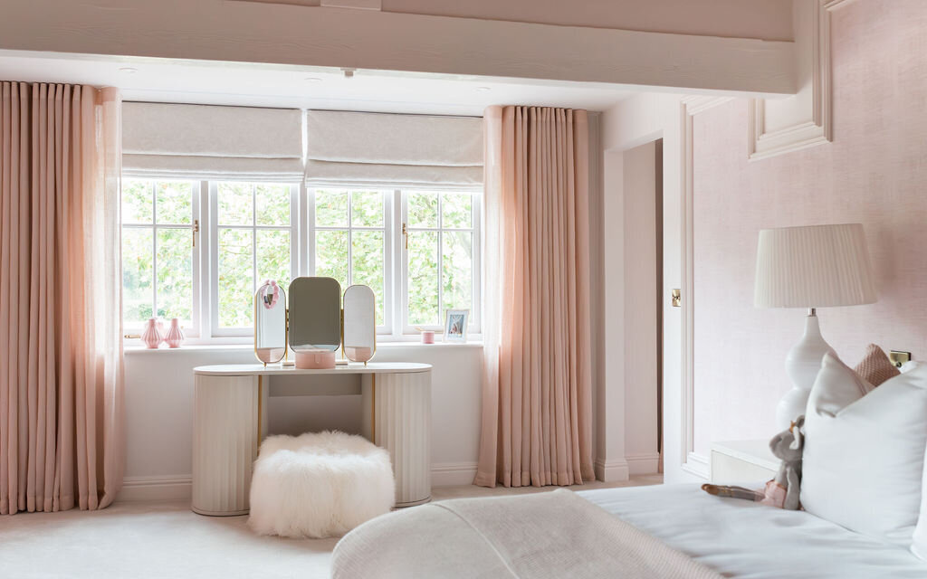 A cozy bedroom with soft pastel decor. The bed is dressed in white and pink linens and fluffy pillows. At the end of the room, a window with pink curtains brings in natural light, and beneath it is a vanity table with a mirror and decorative items.