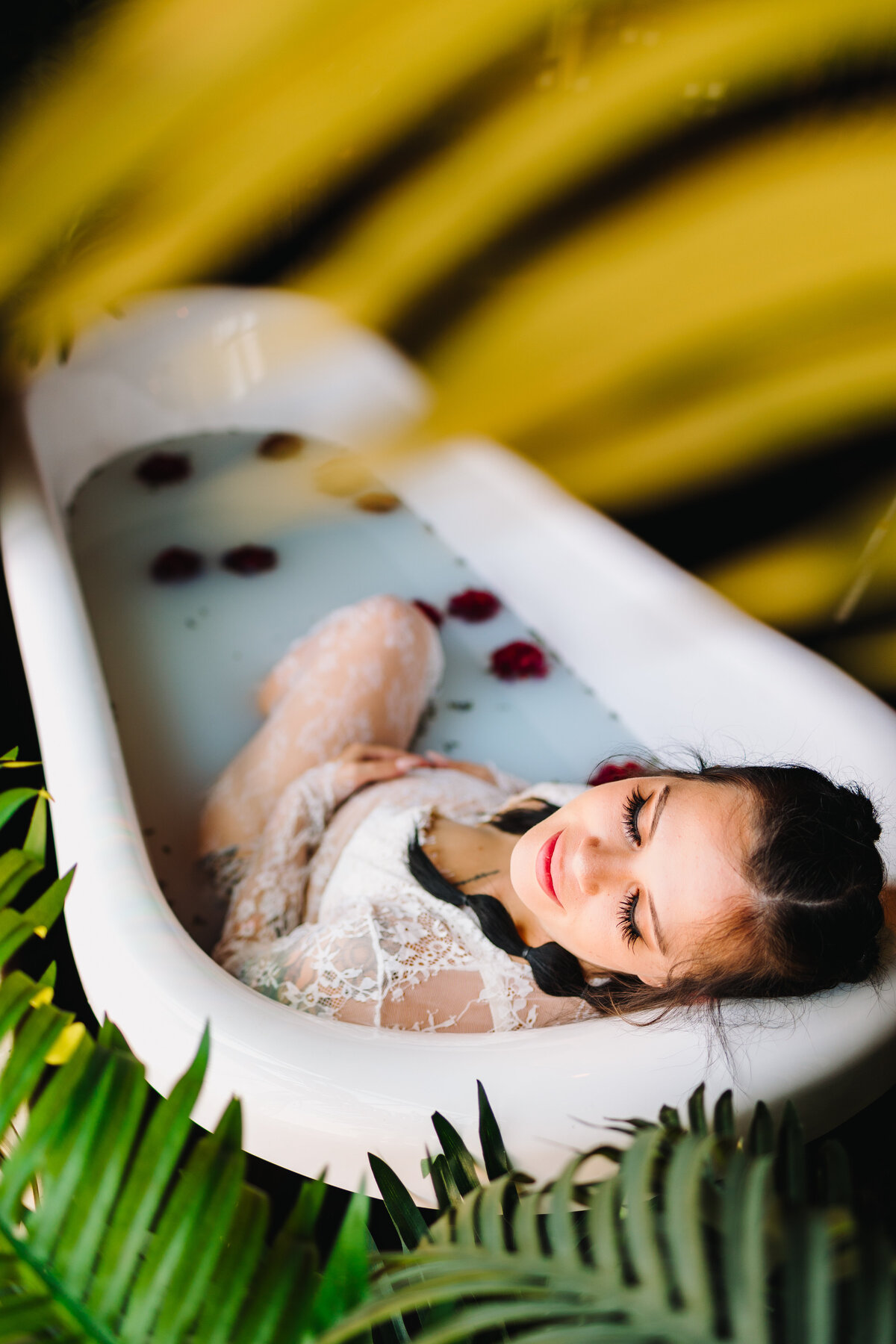 Albuquerque maternity photographer captures a serene moment of a beautiful future mother in a bath filled with red roses. Dressed in a flowing white long dress, she closes her eyes, embodying tranquility.