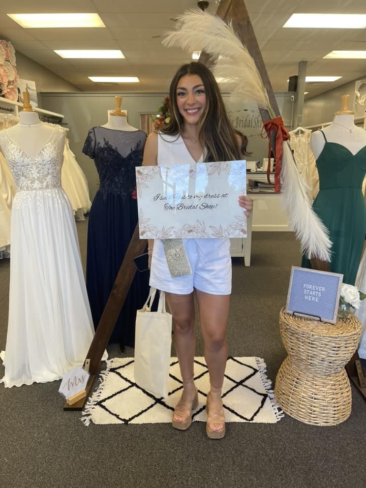 Woman holding a sign announcing her bridal dress purchase inside a bridal shop.