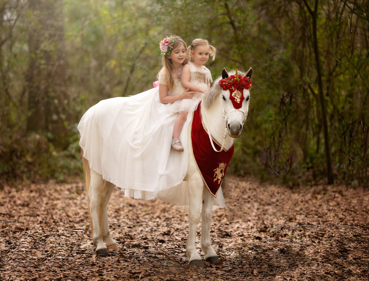 Sisters ride a Welsh pony dressed as a unicorn.  The horse has a red velvet collar and a gold unicorn horn.  The girls are wearing white dresses.  They are in a forest and there are lots of brown leaves on the ground.
