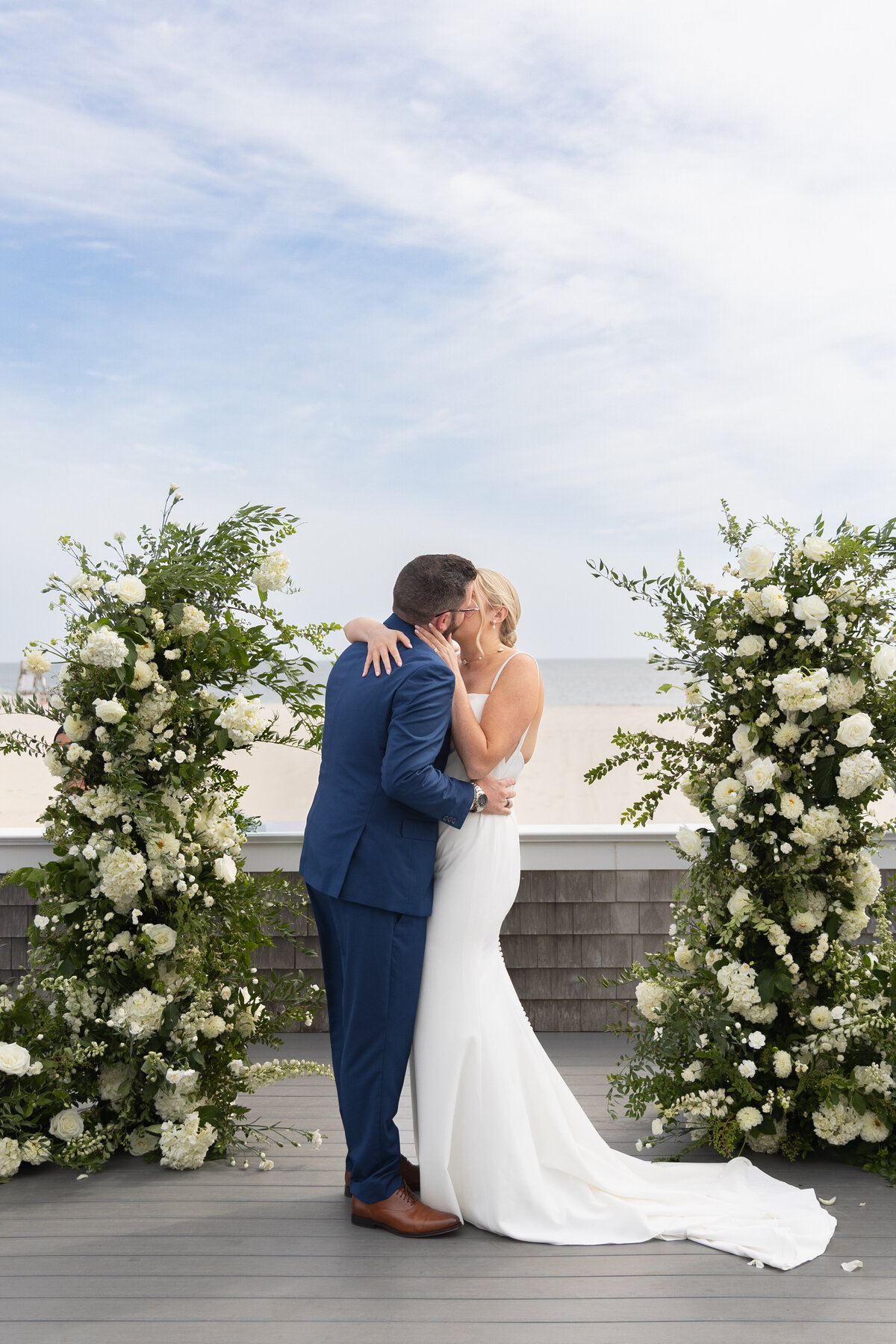 Elopement Wedding Packages Rhode Island | Kelsey Sheehan Photography Timeless Rhode Island Weddings | A bride in a white dress and a groom in a blue suit share a kiss in an outdoor ceremony. They're flanked by lush floral arrangements under a clear sky.