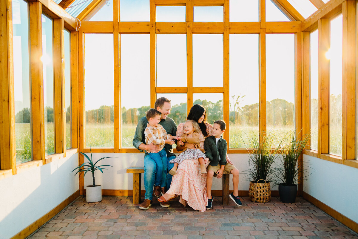 A beautiful family is taking a mini photo session. They are sitting on a wooden chair and all around them are windows.  The woman is wearing a pink dress and the children are dressed in vintage costumes.