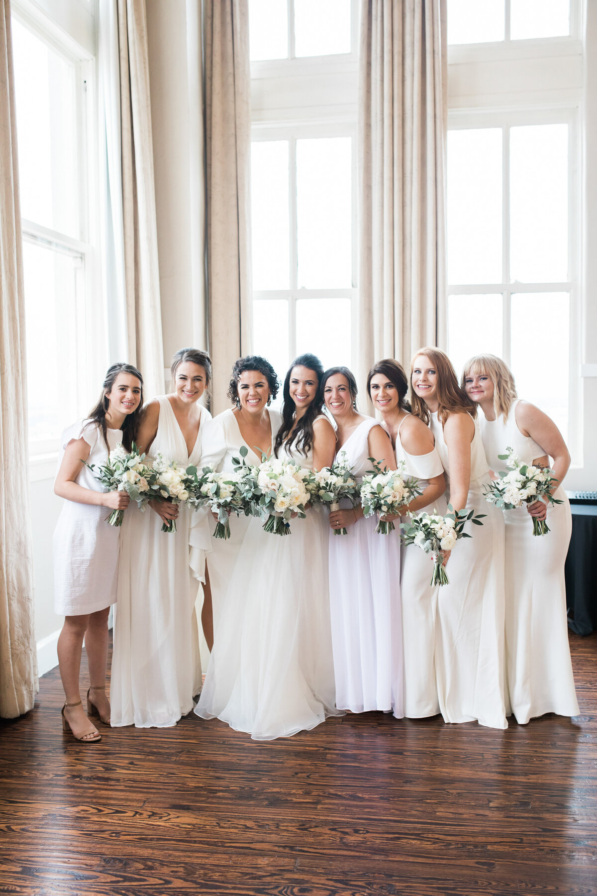 A portrait of a bride and her bridesmaids before her wedding ceremony at the Room on Main in Dallas, Texas.  Everyone, including the bride, is wearing white dresses and holding bouquets of white flowers. They are all smiling and standing very close to each other.