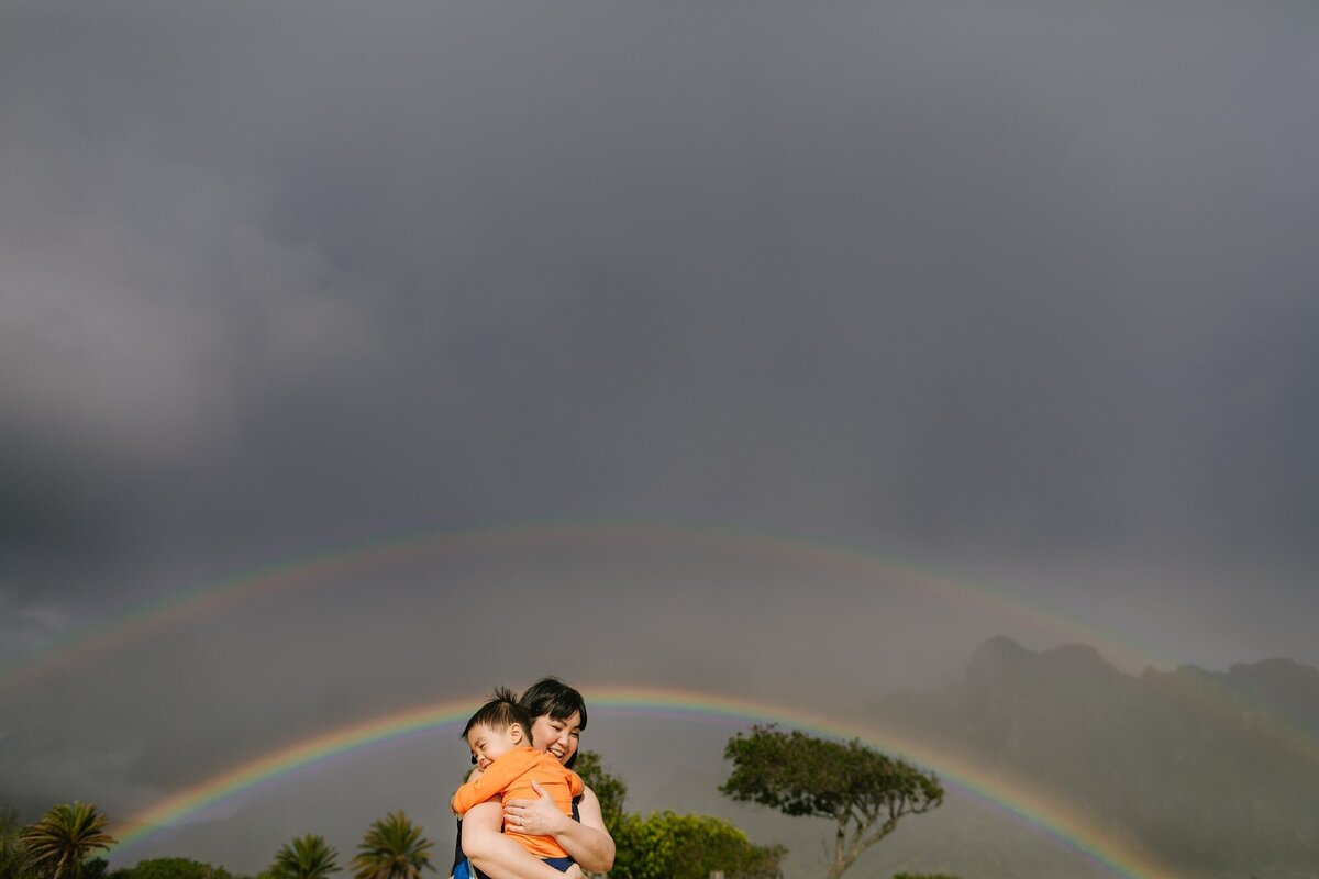 mom and son share a hug with a rainbow behind them at kualoa beach