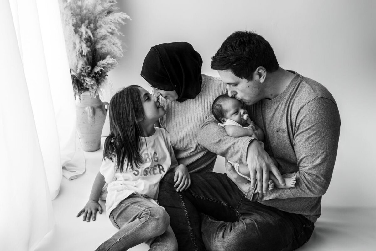 a loving family: a father kissing his newborn, a mother in a hijab sharing a sweet moment with her older daughter.