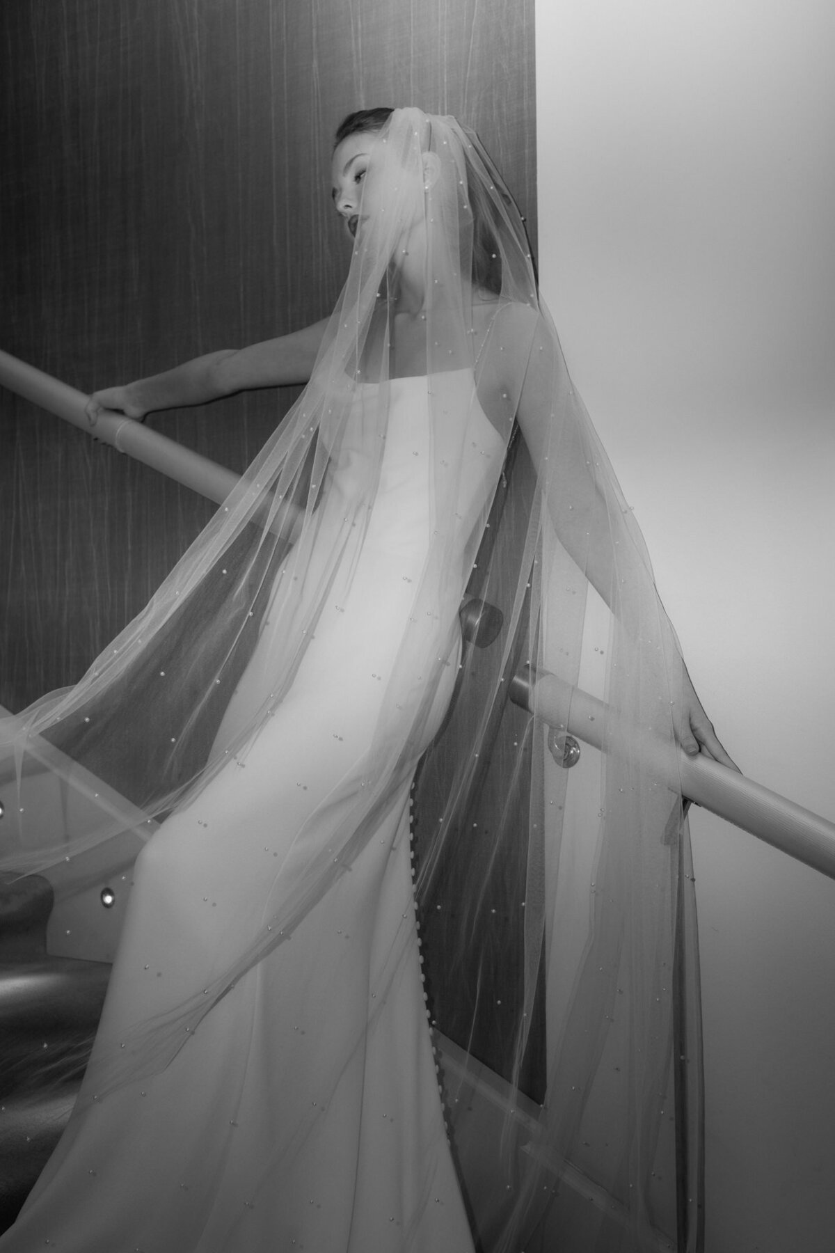 Den langen Schleier mit den kleinen Perlen präsentierend lehnt das Brautmodel an einem Treppengeländer.