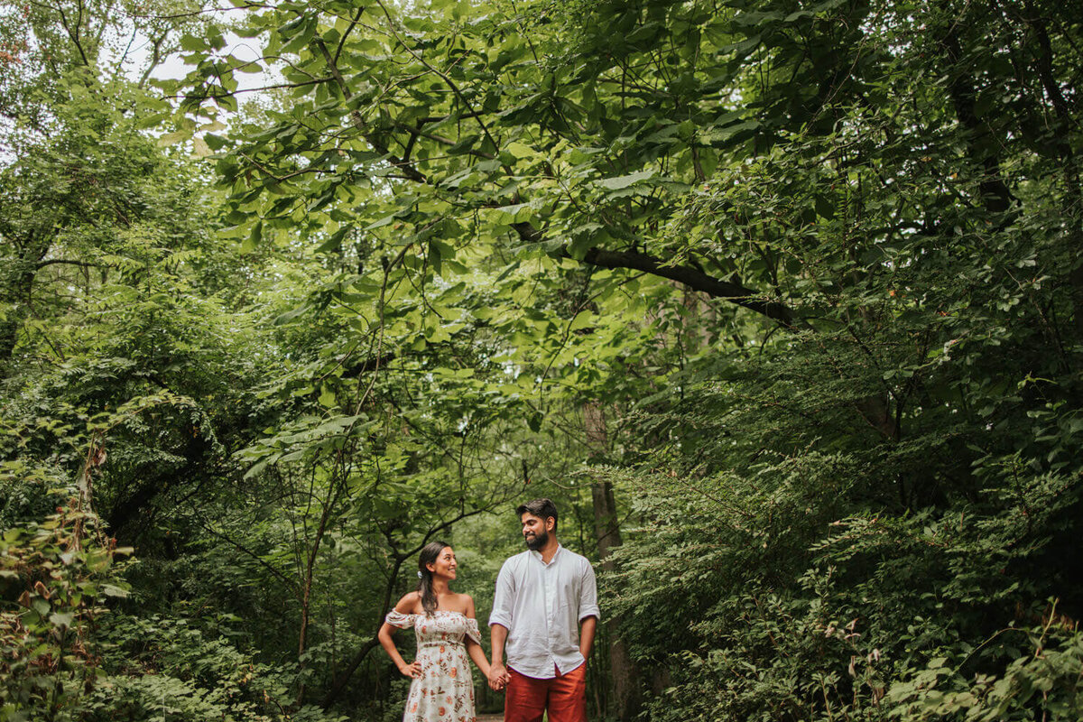A couple strolls hand in hand through a lush, green forest in Sayen House and Garden in Hamilton Square, NJ, sharing a warm and affectionate gaze.