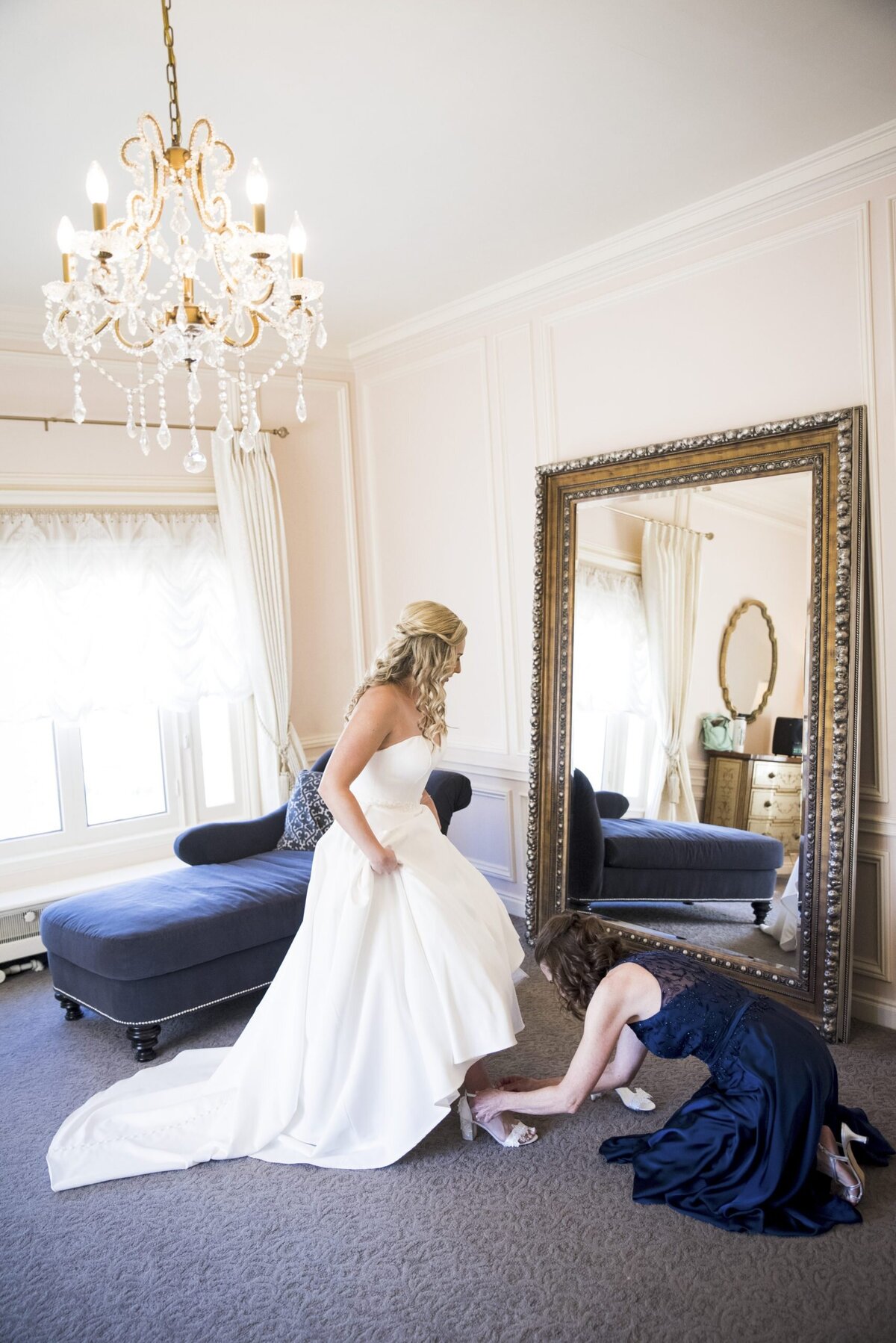 A bride looks in an ornate mirror as one of her bridesmaids helps her fasten her shoe.