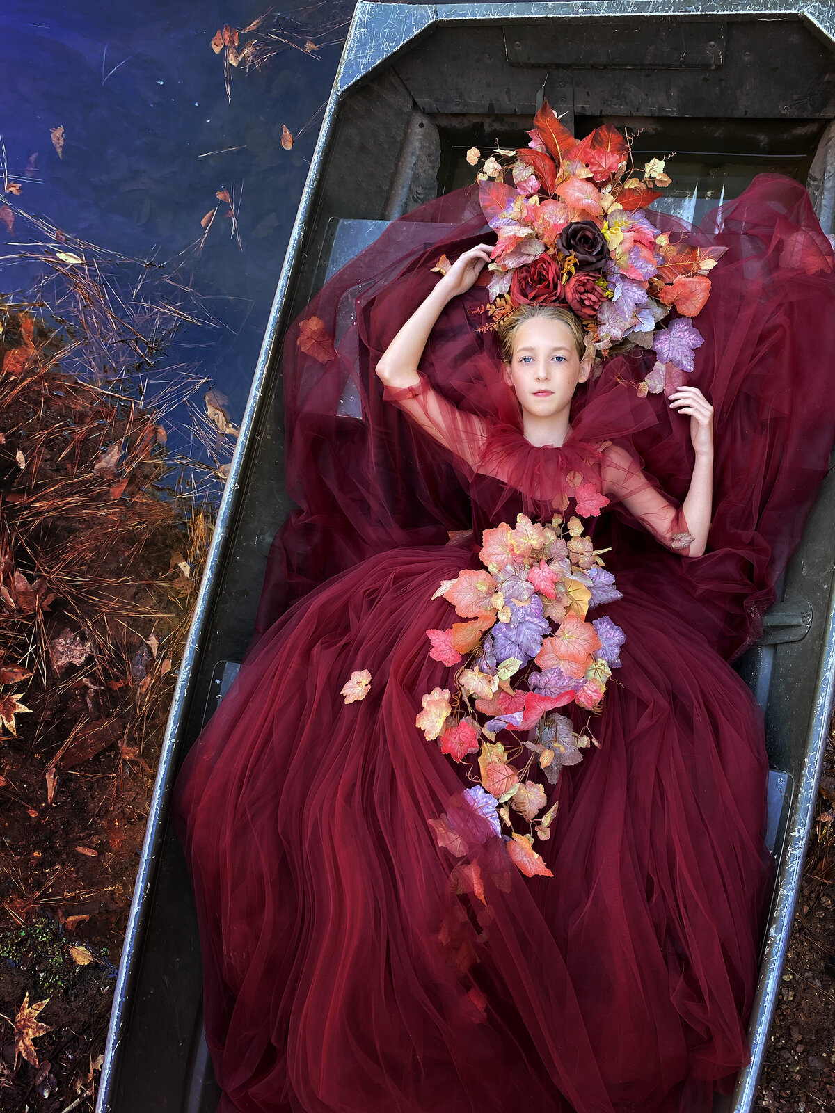 A young girl lays in a boat surrounded by vibrant fall foliage, wearing a rich burgundy tulle gown adorned with autumn leaves. Her elaborate floral crown enhances the scene's ethereal feel, reflecting the warmth of the season. The serene water and surrounding nature complete the composition.