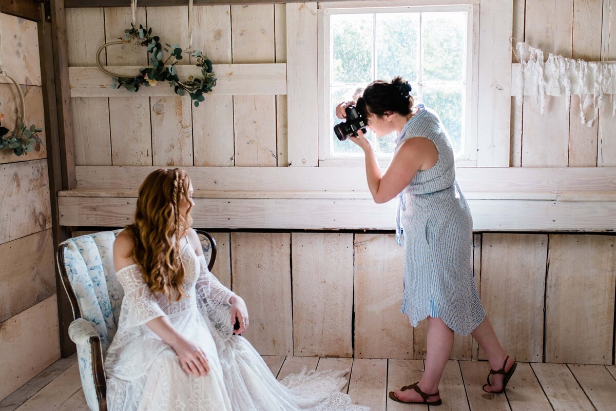 Image of woman in a blue sundress taking a photo of a red headed bride in a wedding gown sitting in a chair