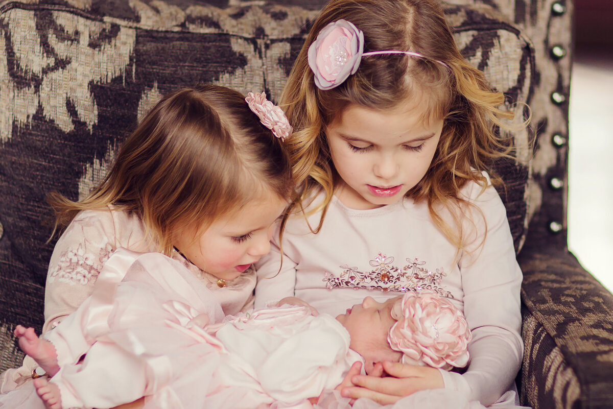 Older sisters are sitting in a chair holding their new sister, looking at her adoringly. All are wearing pink.