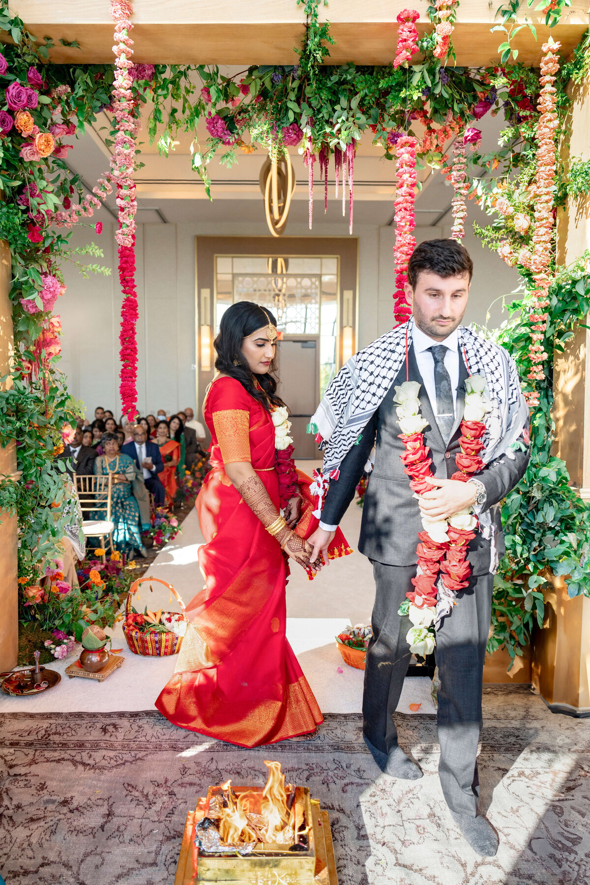 A couple participates in a wedding ceremony. The bride, in a red saree, holds hands with the groom, who wears a suit and shawl. They walk around a small fire, surrounded by colorful floral decorations and guests in the background.