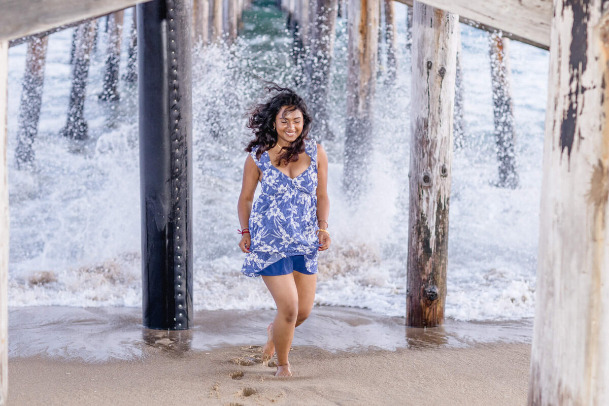 A woman in a blue and white dress runs along a sandy beach beneath a wooden pier as waves crash dramatically against the pillars behind her. She is smiling and appears joyful.