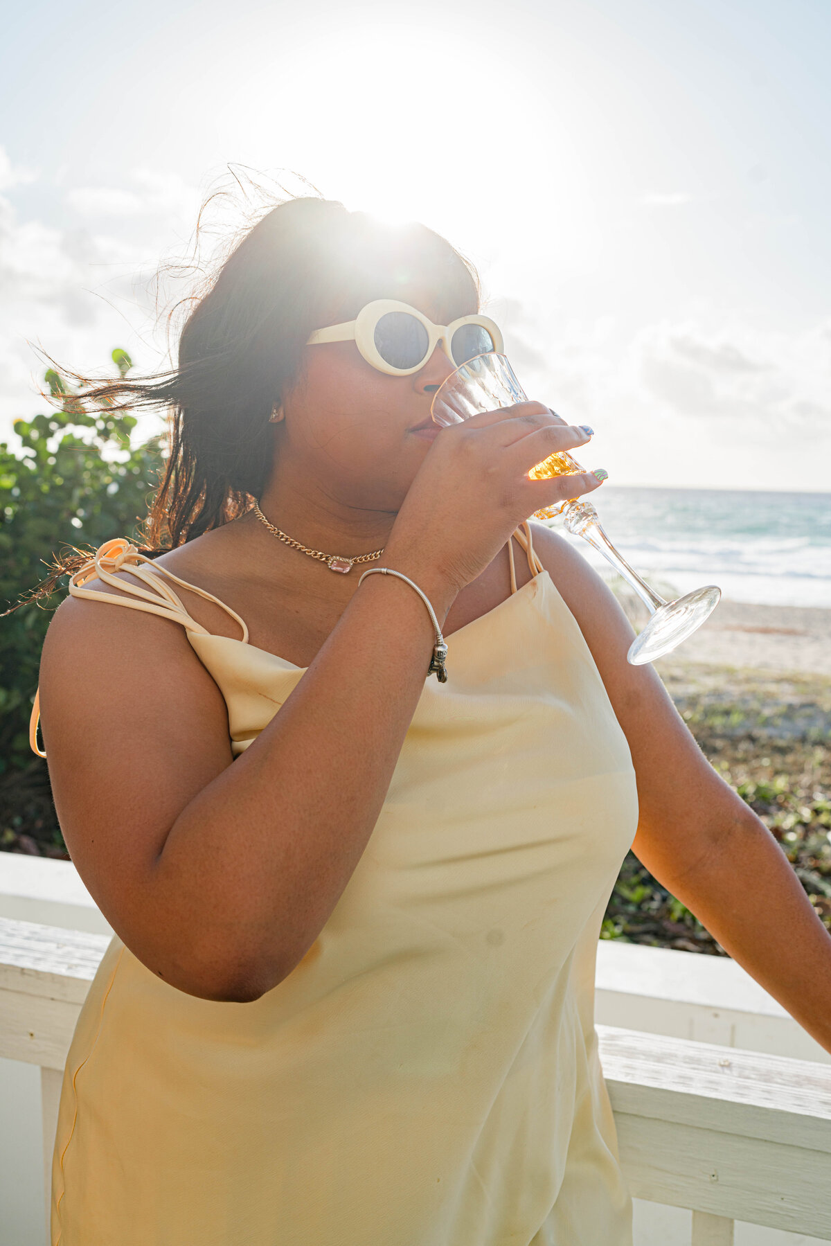 girl in yellow dress with white sunglasses on the beach drinking champagne from a crystal glass