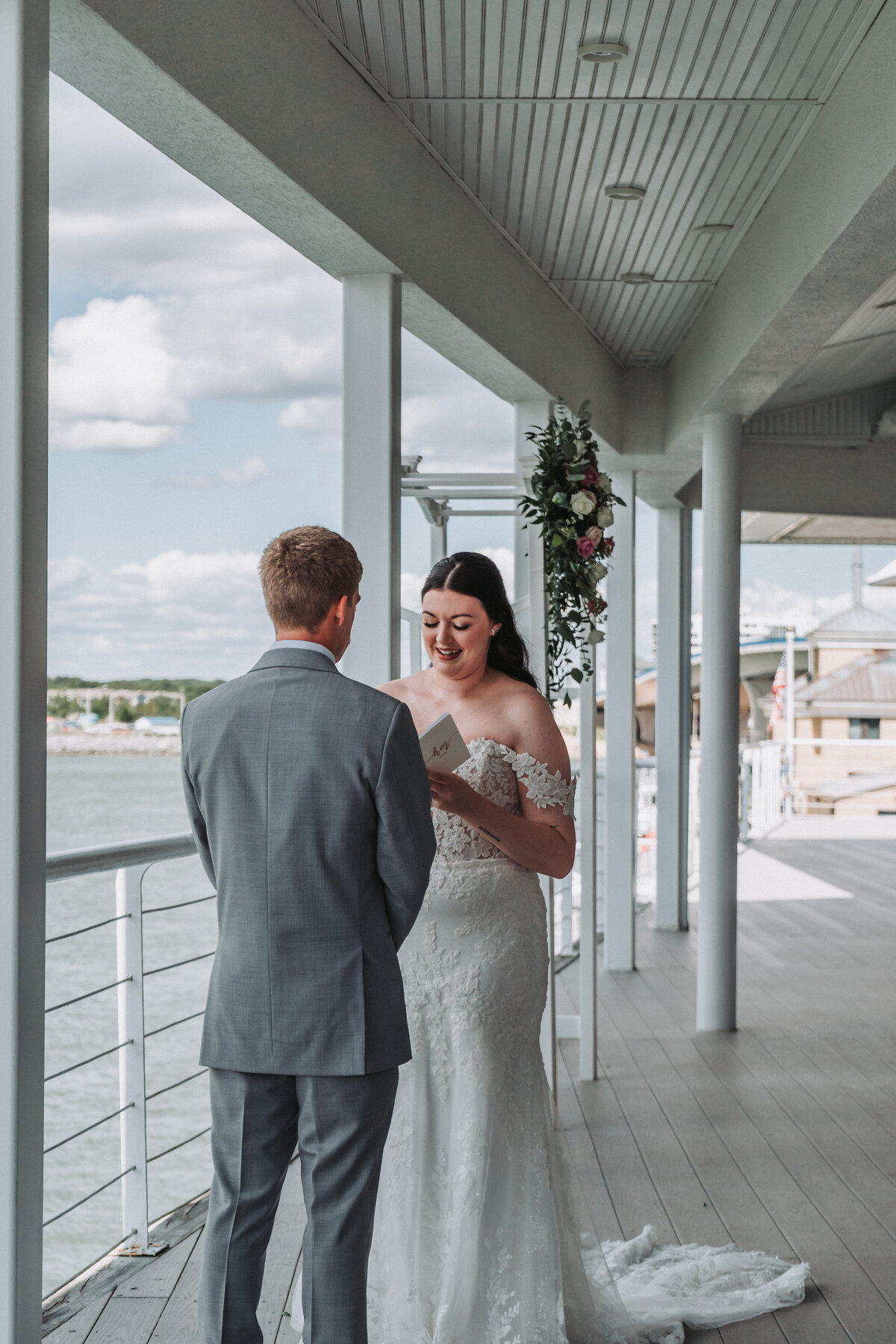 A beautiful bride, in an off-the-shoulder lace gown, reads her vows to the groom during their outdoor waterfront wedding ceremony at the Lesner Inn in Virginia Beach, captured by Quinn Photography LLC.
