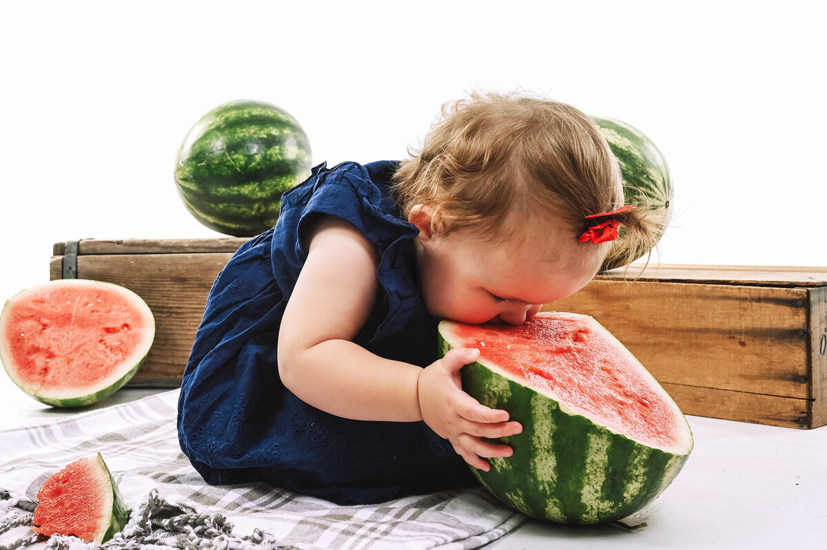 Toddler sitting on a blanket on a white background bends to take bite of watermelon