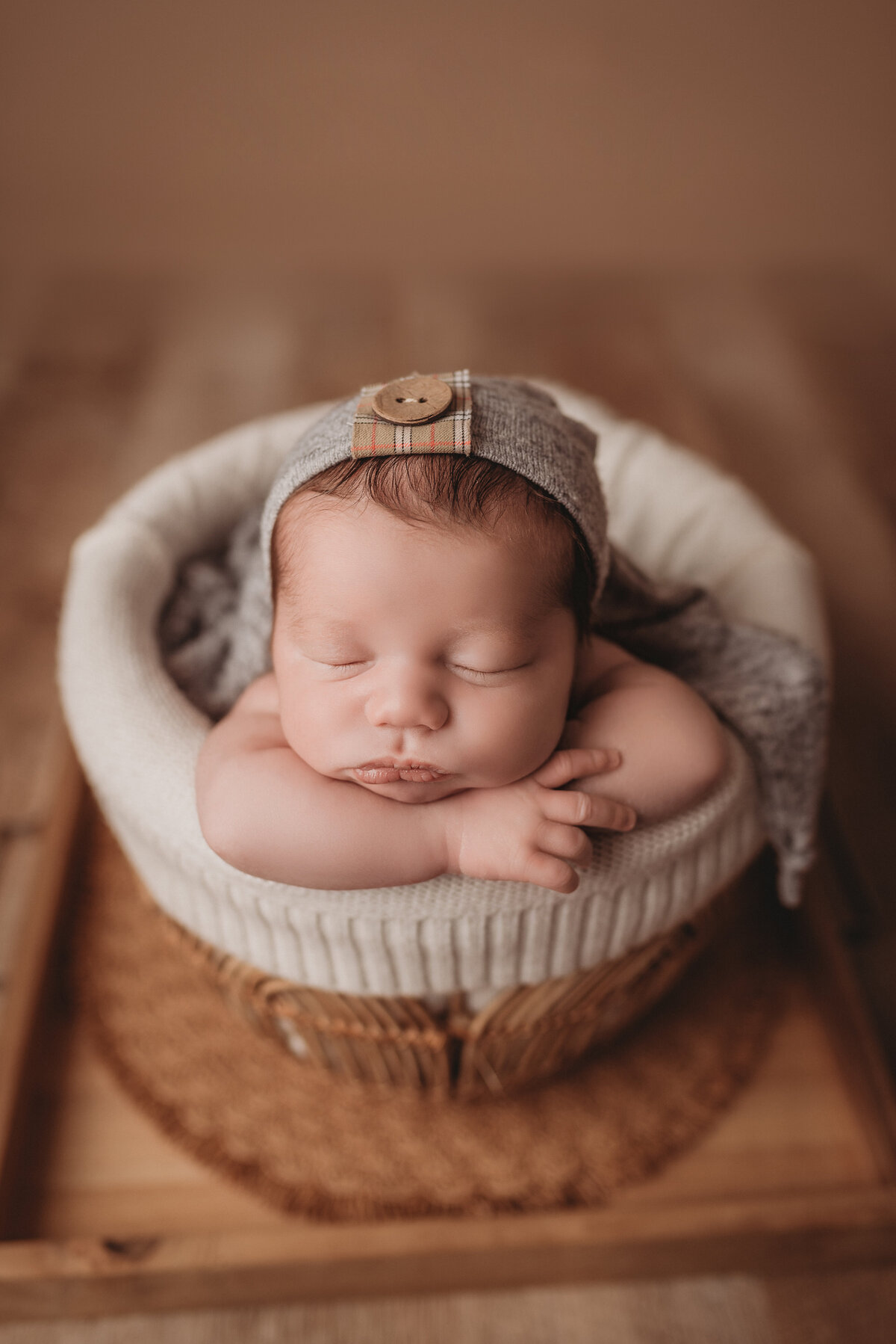 Newborn baby boy sitting in a rattan basket with hands under chin, wearing a gray sleepy cap on wooden backdrop