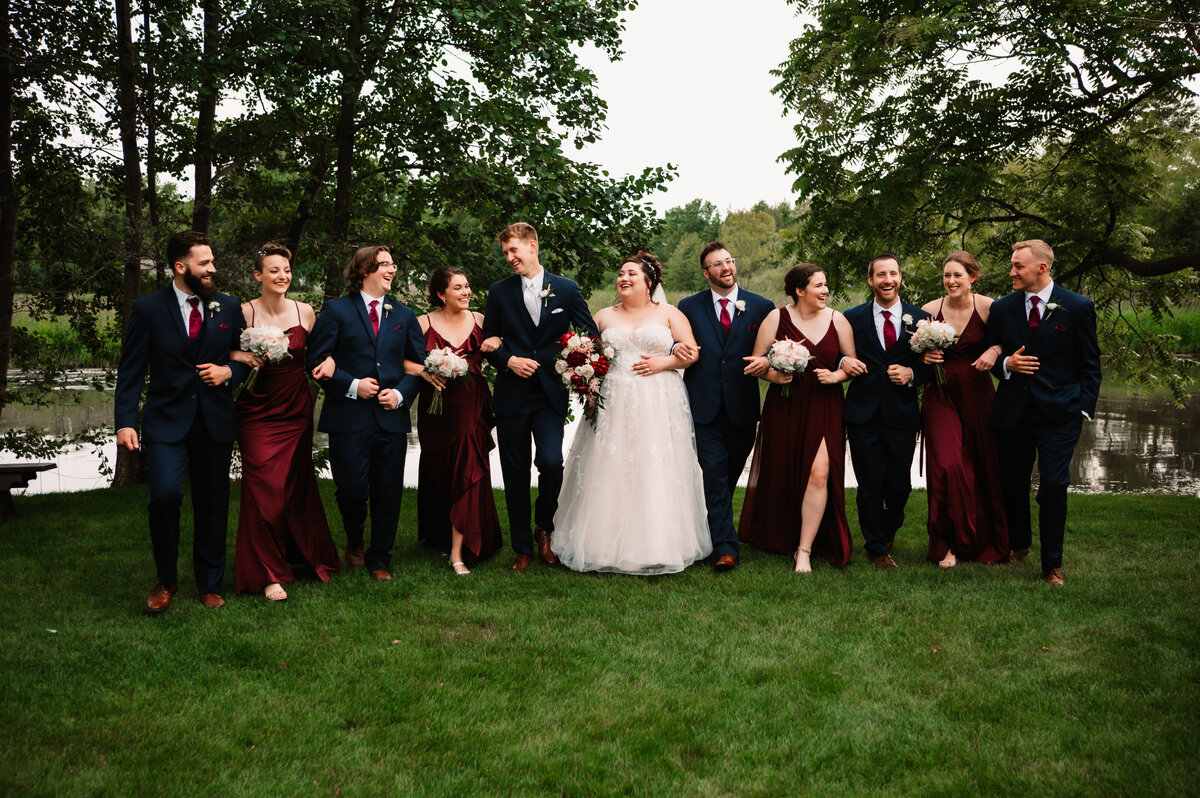 Newlyweds walk laughing with their wedding party on a lawn by a pond