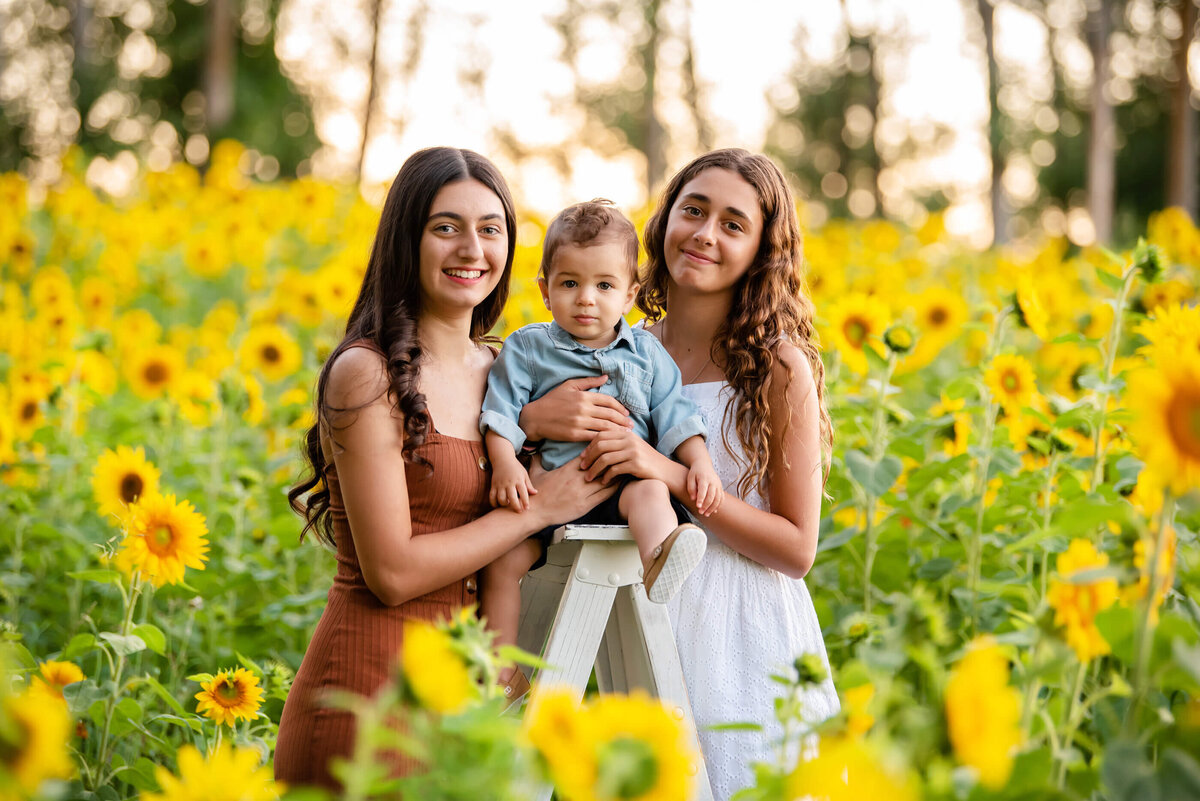 Teen Sisters with their toddler brother sitting on a white ladder in a sunflower field for their summer outdoor family photography session in Burlington, On