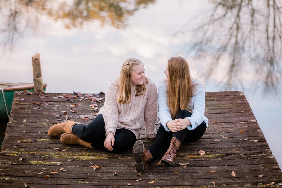 Two sisters sitting on a dock with the sky and trees reflected in the water behind them as they share a secret.