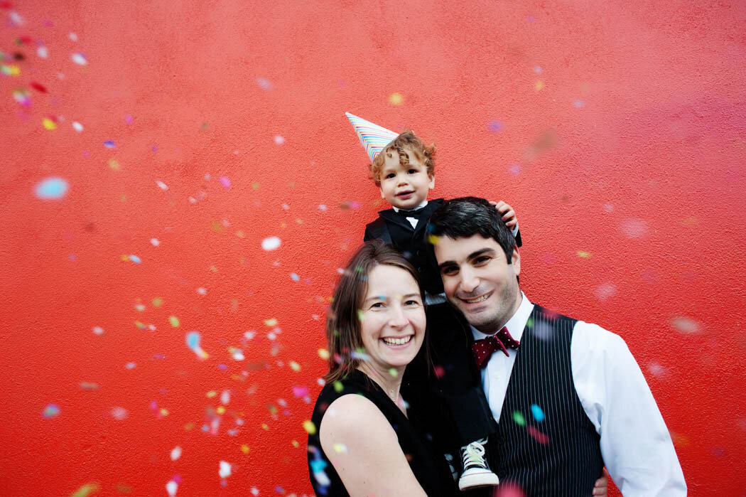 Smiling mom and dad with young son on their shoulders with confetti, in front of a vibrant red background.