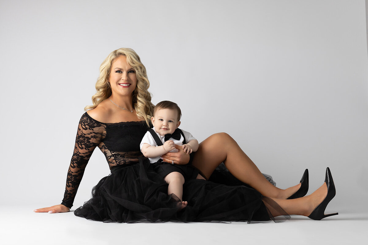 A glamorous mom in a black lace gown sits with her baby boy dressed in a tuxedo romper, both smiling during a studio milestone session with a Dallas milestone photographer.