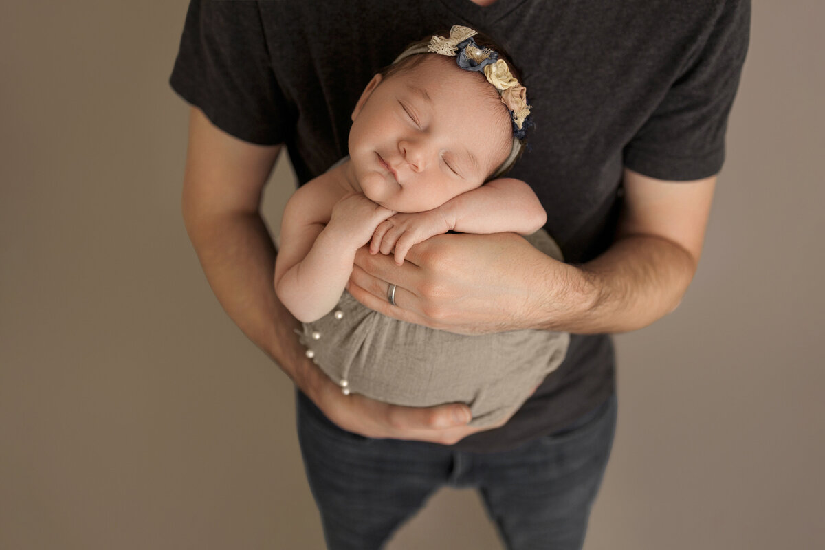 Father holding his sleeping newborn baby wrapped in a grey blanket, baby's head resting on his arms, posing against a neutral beige background.