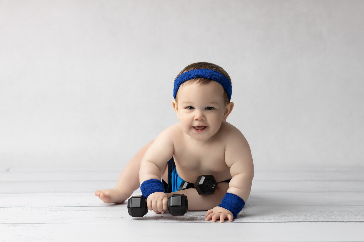 Smiling baby boy crawling with toy dumbbells on a white floor, wearing a blue headband and wristbands, captured by a Dallas milestone photographer.