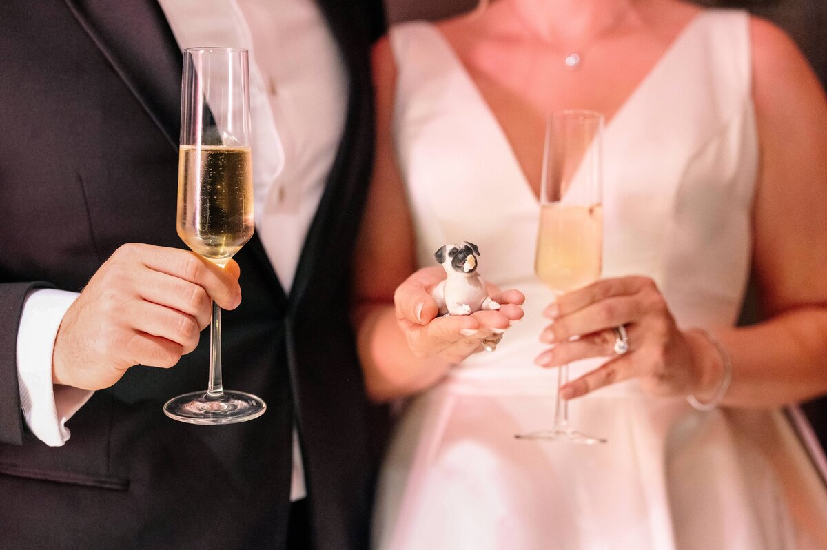 A couple in formal attire, with the man in a tuxedo and the woman in a white dress, hold champagne flutes. The woman holds a small figurine of a dog in her other hand.