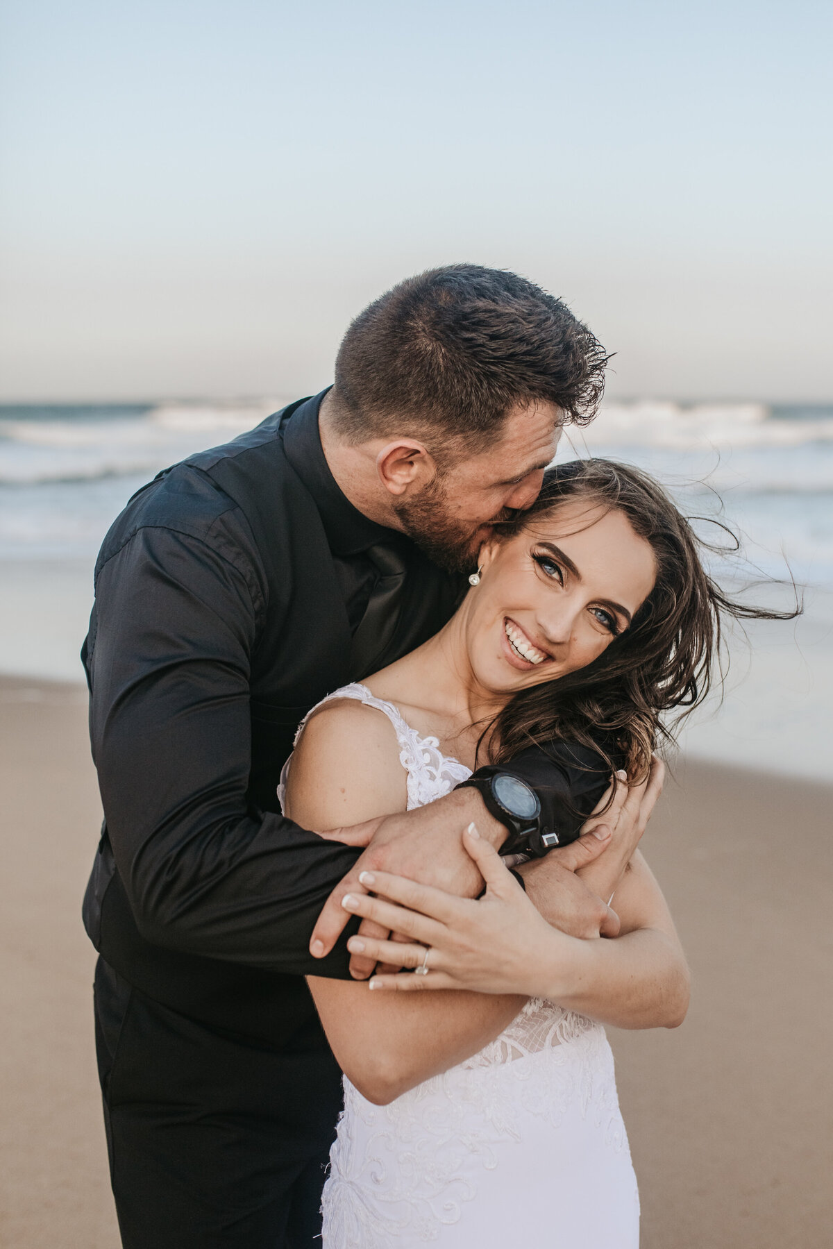 The groom wraps his arms around the bride while kissing her on her head on the beach. 