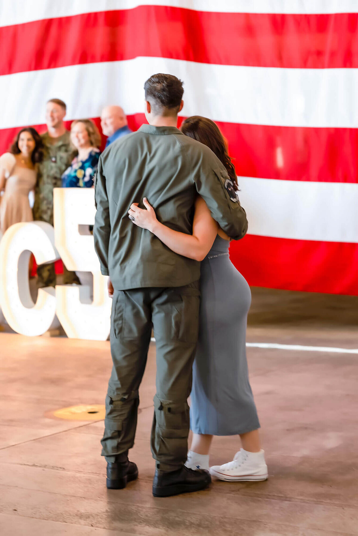 A couple snuggles close at a military homecoming. They watch as other families get their pictures taken with the American flag and squadron sign.