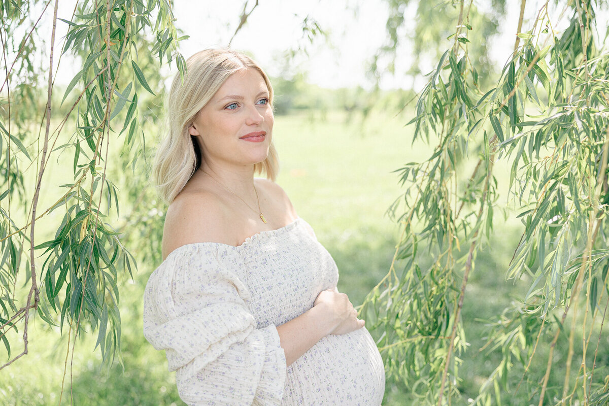 Pregnant mother in floral nothing fits but dress stands among a willow tree while holding her baby bump and looking into the distance, Indianapolis Spring Minis