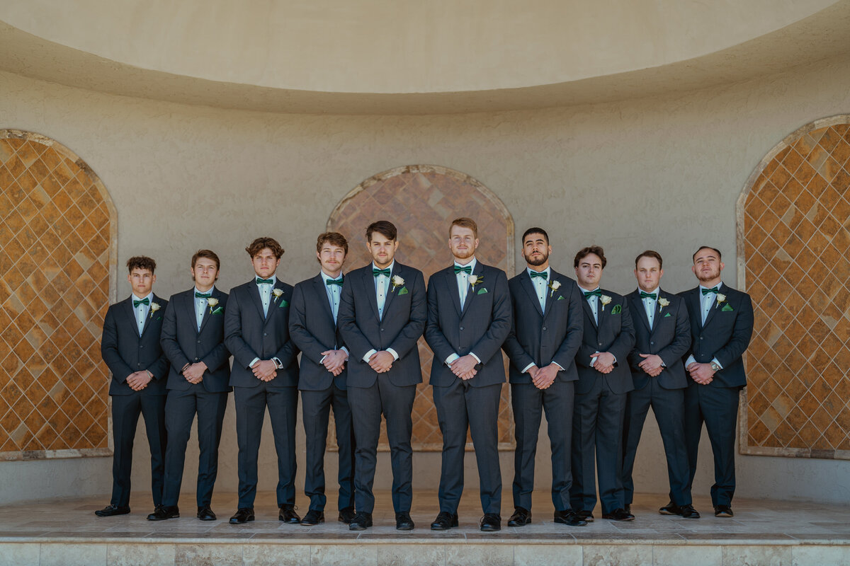 Formal group photo of groomsmen in matching suits with green bow ties, posing at D'Vine Grace wedding venue. The elegant backdrop features architectural details, adding a touch of sophistication to the image.