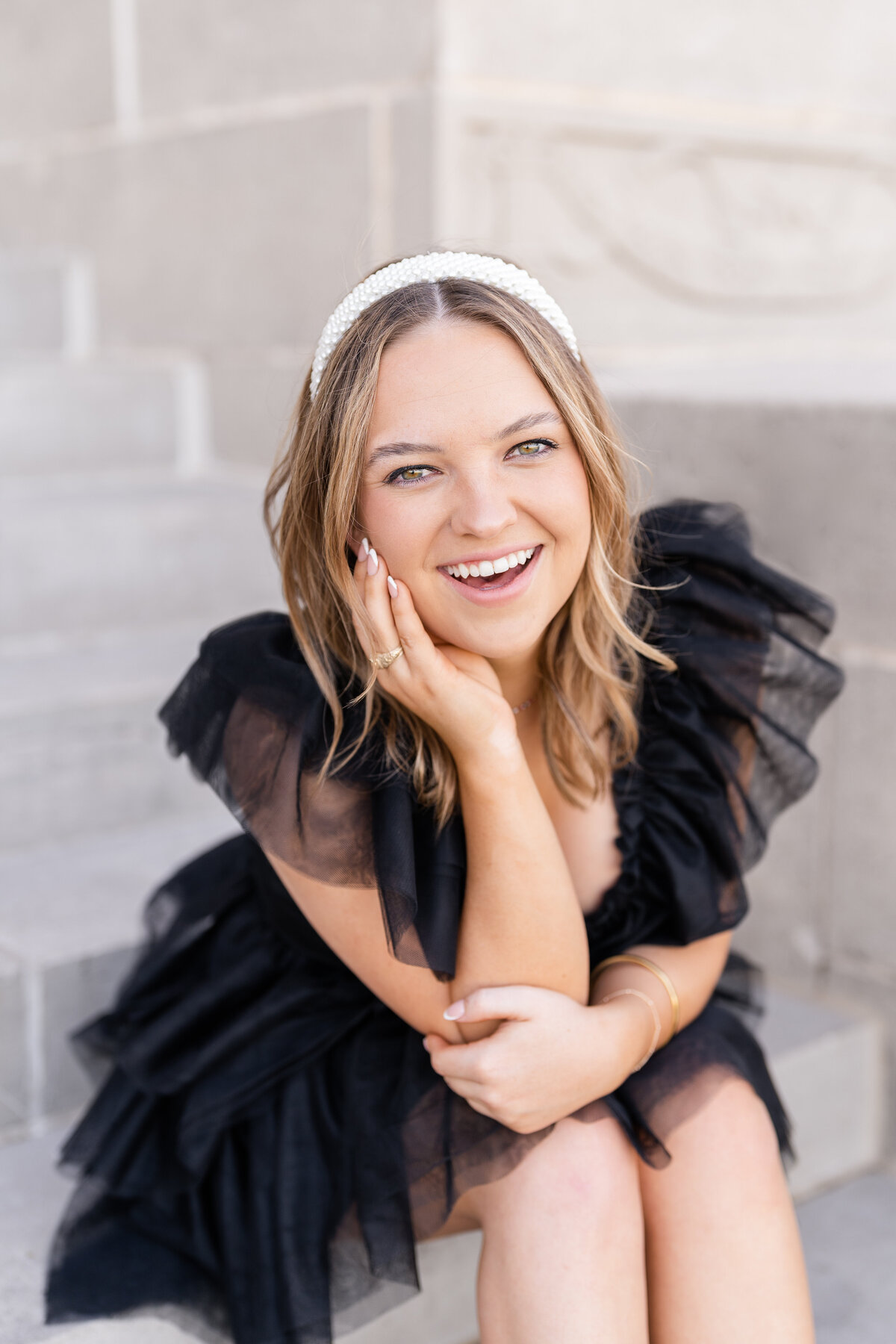 Texas A&M senior girl sitting on Administration Building stairs wearing a fluffy black dress and smiling with pearl headband and hand on cheek