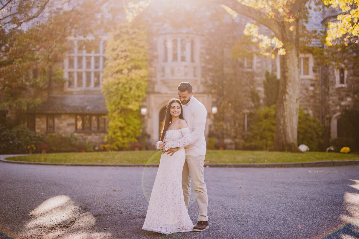 An engaged couple in white clothes standing in front of the skylands manor at sunset at ringwood state park in nj