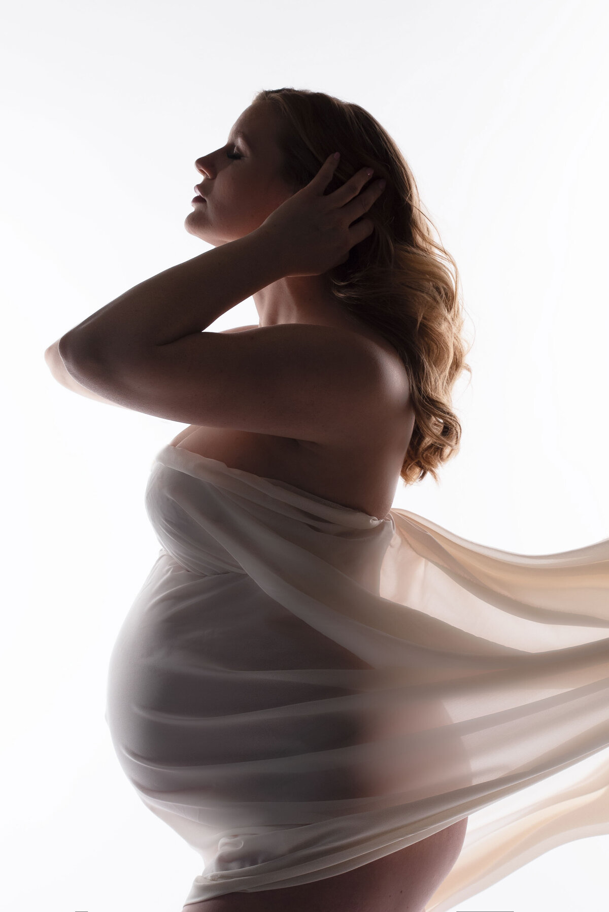 Pregnant woman wrapped in white chiffon fabric standing turned to the side with hand behind head