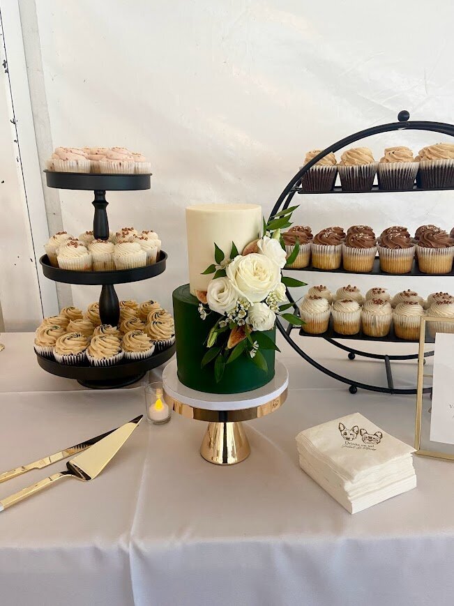A wedding dessert display shows a two tier cutting cake with a bottom emerald green tier and white top tier, with a cluster of white flowers and fresh fig accents. Behind the cake are two large black stands filled with different types of cupcakes.