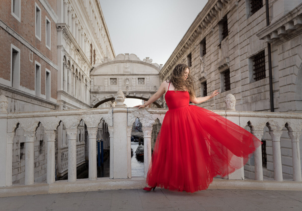 Woman in a red tulle dress with the skirt flowing out standing on a bridge at sunrise in St Marks Square, Venice. Taken by beauty photographer Claire Howes, Norwich.