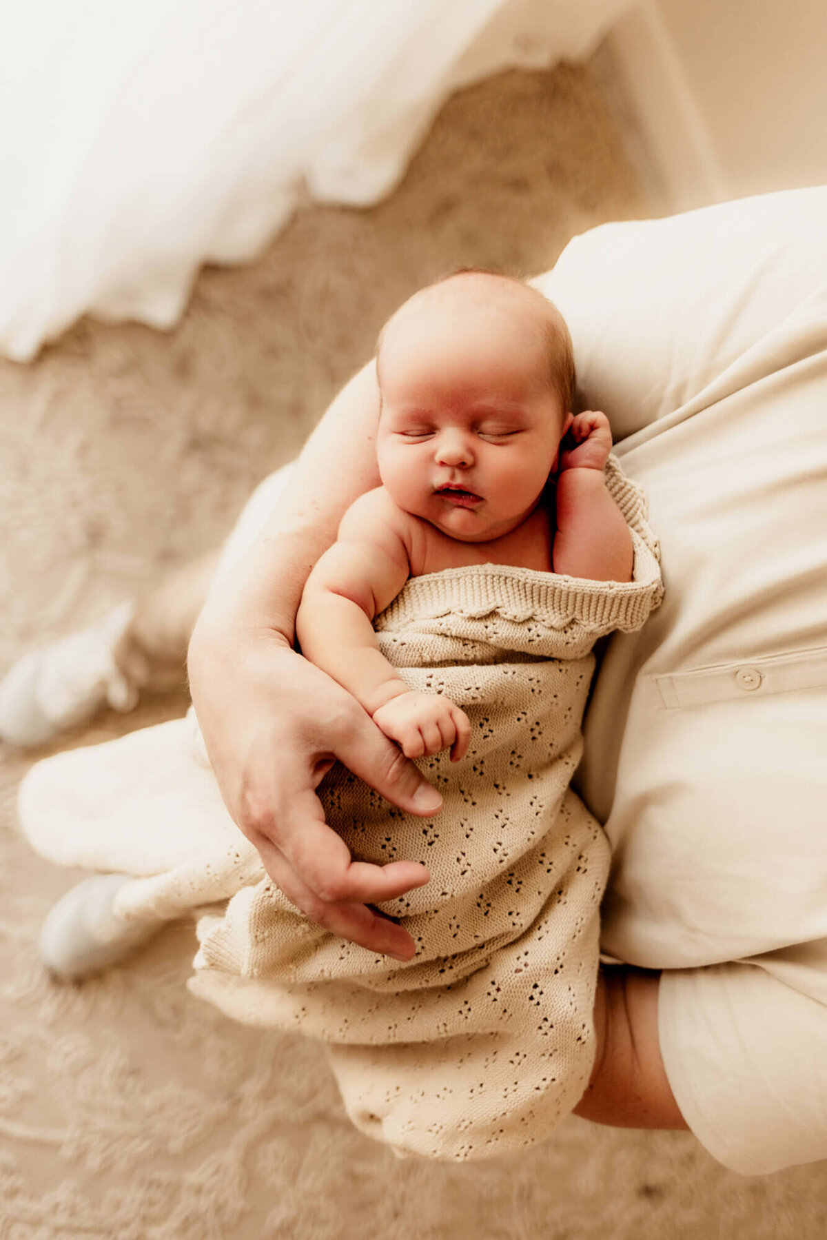 Baby boy swaddled in a beige blanket sleeping in his father's arms. 