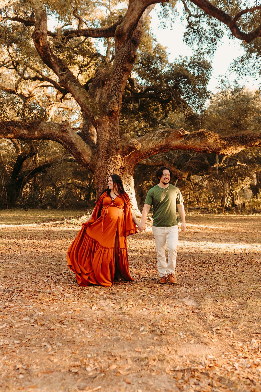 Dayton maternity photography – expecting couple walking hand in hand in outdoor oak grove, burnt orange maternity gown