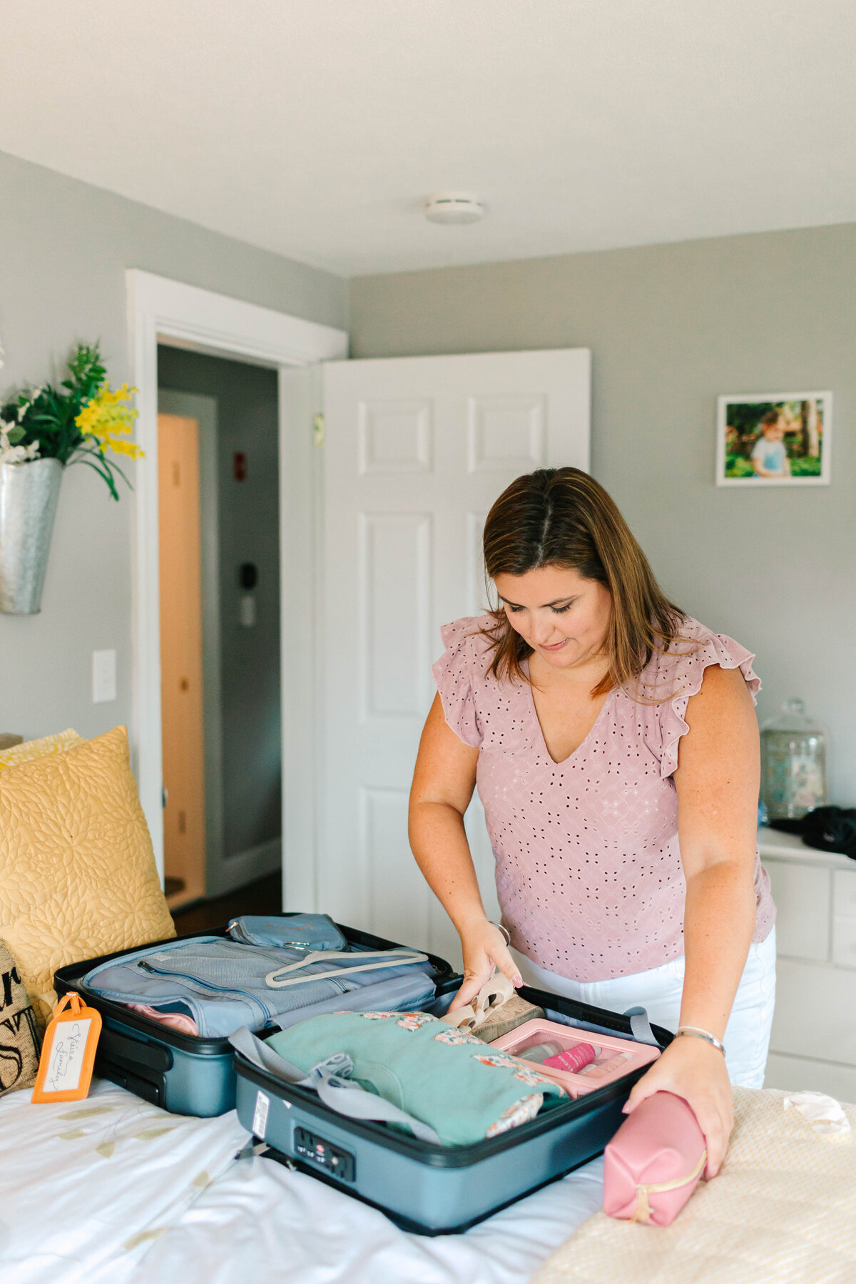 A woman in a ruffled mauve top packs a neatly organized suitcase on a bed in a cozy, well-decorated bedroom. A pink toiletry bag rests beside her as she arranges folded clothes and travel essentials.