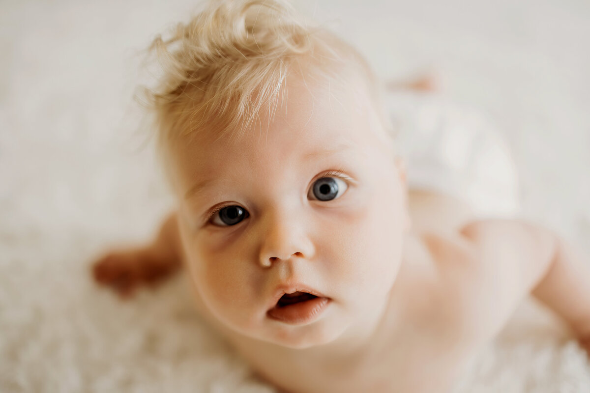 A happy six month old boy lays on his belly on a white rug in a white diaper cover for his milestone photos in Denver. 