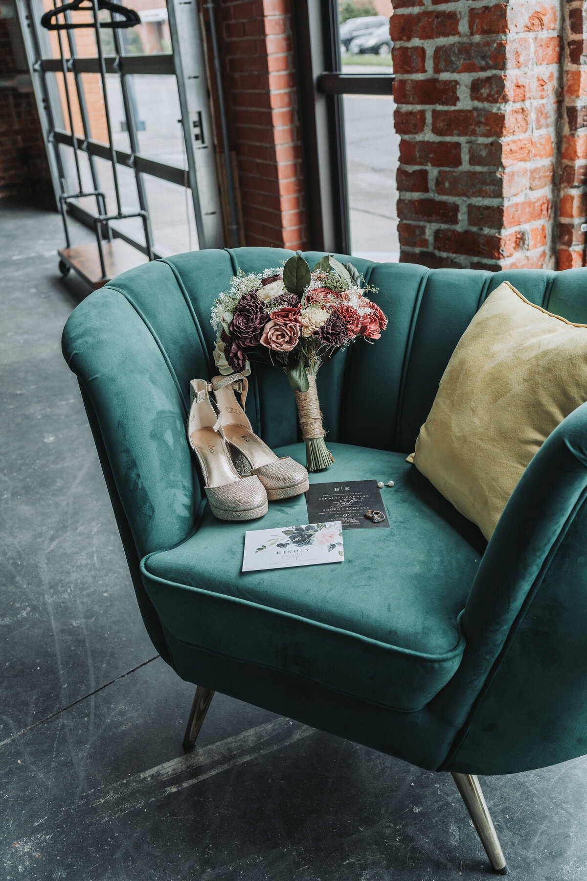A beautiful teal velvet armchair holding a rustic wedding bouquet, sparkly bridal heels, and elegant wedding invitations, set against a brick wall backdrop.