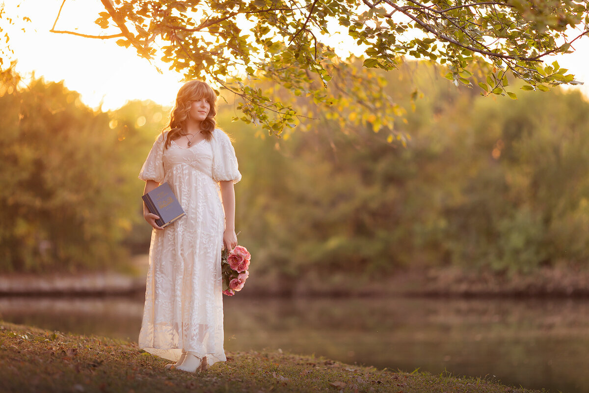 Dallas senior photographer captures a young woman in a white dress holding a book and a bouquet of pink roses during sunset by the water.